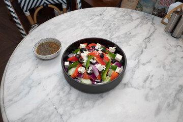 A close-up shot of a Greek salad in a black bowl, along with a small bowl of dressing on a marble table