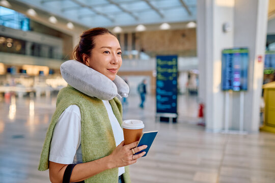 Young Asian woman wearing a neck pillow, holding passport and coffee, waiting for flight in a modern airport terminal