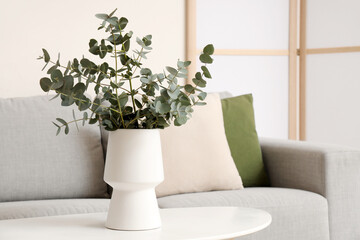 Vase with fresh eucalyptus branches on table near grey sofa and folding screen in living room