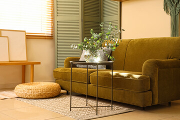 Obraz premium Glass vase with fresh eucalyptus branches on table near green sofa and folding screen in living room