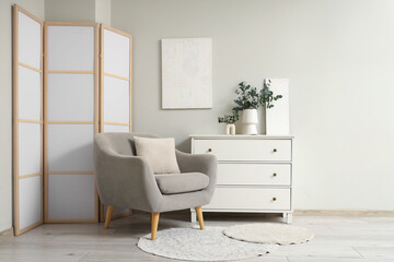 Vases with fresh eucalyptus branches on white chest of drawers near grey armchair and folding screen in living room