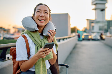 Young woman traveler smiling, holding smartphone and wearing a neck pillow while waiting for departure at the airport