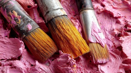 Three paintbrushes are sitting on a pink surface. The brushes are old and worn, with some paint still on them. The pink surface is likely a canvas or a piece of paper