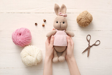 Female hands with knitted bunny toy, yarn and scissors on white wooden background
