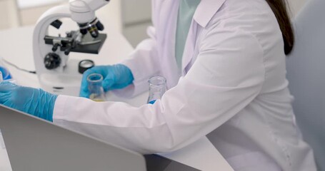 Close up of Asian schoolgirl wearing gloves and labcoat holding beakers filled with yellow and blue chemical solutions beside microscope and laptop during science experiment in school laboratory