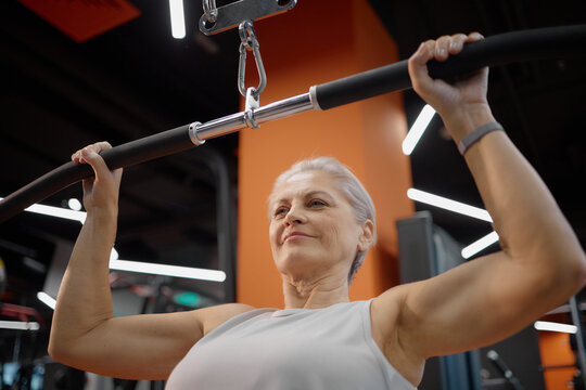 Senior Caucasian woman exercising on lat pulldown machine in gym, gripping bar with both hands, looking focused while performing strength training workout, visible upper body muscles