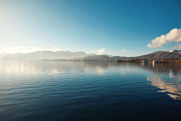 Serene lake landscape with calm waters reflecting mountains and clouds under a clear blue sky, showcasing natural beauty and tranquility