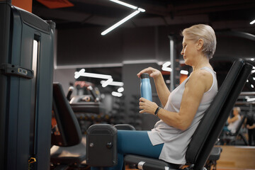 Senior Caucasian woman exercising on leg press machine in gym, holding water bottle and taking break during workout, short gray hair visible, focused on fitness routine