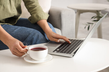 Young woman with cup of tea and laptop near coffee table at home, closeup