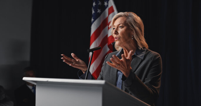 Politician, speech and woman with American flag by podium for election, debate and voting campaign. Government, candidate and mature person on stage for press conference, political policy and rally - Powered by Adobe