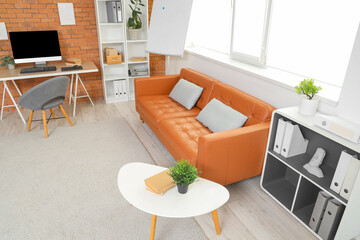 Interior of modern office with shelving units, workspace and cozy orange sofa near window