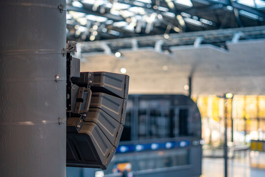 Public address speaker array mounted on metal column inside modern transportation building, part of passenger voice announcement and audio communication system