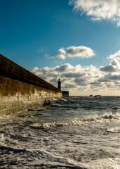 A lighthouse stands on a stone wall as waves crash against it under a cloudy sky.