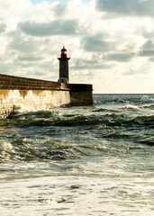 A lighthouse stands on a stone pier as waves crash against it.