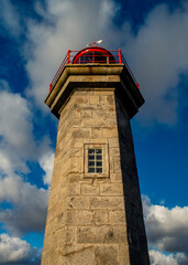 A tall stone lighthouse with a red railing and a weather vane on top, set against a blue sky with clouds.