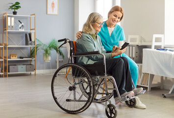 Healthcare worker assists woman in wheelchair by explaining medical app on her smartphone. Female...