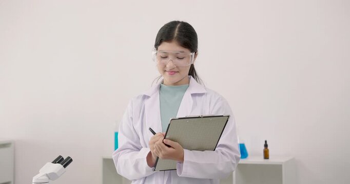 Asian young girl smiling while holding clipboard during science class in school laboratory, wearing labcoat and safety goggles with colorful chemical liquids and equipment in background