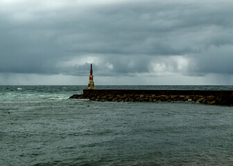 A lighthouse stands on a stone pier in the ocean under a cloudy sky.