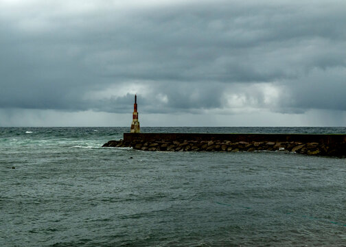 A lighthouse stands on a stone pier in the ocean under a cloudy sky.