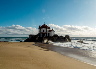 A chapel on a rocky outcrop on the beach, with waves crashing around it.