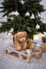 Brown vintage Teddy bear sitting on wooden sledge with Christmas tree and Christmas lights and presents in the background.	
