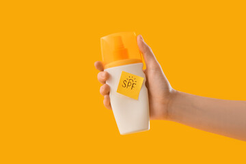 Woman holding bottle of sunscreen cream and sticky note with word SPF on yellow background