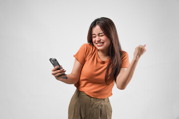 Fototapeta premium Excited young Asian girl raising fists with happy successful expression while holding smartphone.