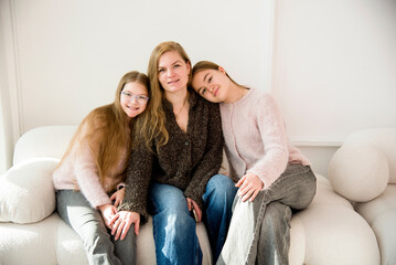 beautiful blonde smiling mother and daughter with long hair sitting on sofa in white studio
