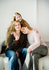 beautiful blonde smiling mother and daughter with long hair sitting on sofa in white studio