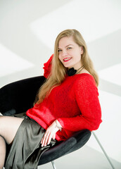 young blonde smiling beautiful lady in red handmade sweater and black skirt and black flower on the neck sitting on a chair