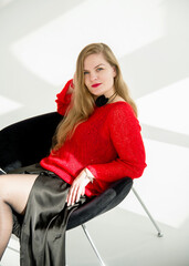 young blonde smiling beautiful lady in red handmade sweater and black skirt and black flower on the neck sitting on a chair