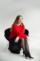 young blonde smiling beautiful lady in red handmade sweater and black skirt and black flower on the neck sitting on a chair