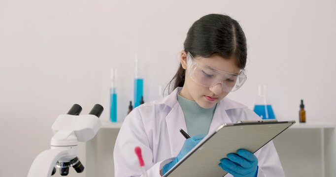 Science student girl checking notes on clipboard with microscope and laboratory glassware on desk wearing goggles and gloves in classroom