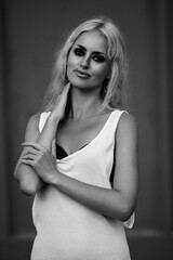 Naklejka premium Young woman with long blonde hair, wearing a stylish white dress, poses confidently with a soft smile, showcasing elegance and beauty in a monochrome portrait