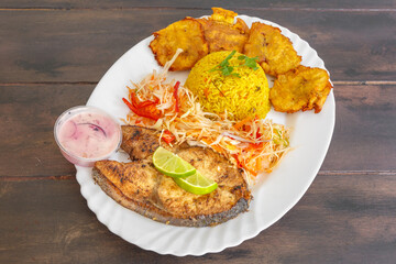 Poisson frit servi avec bananes pesées, pikliz aux poivrons et riz Madras, cuisine haïtienne traditionnelle sur une table en bois.