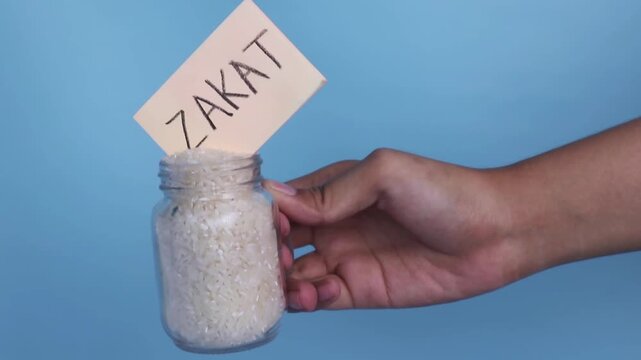A hand holds a small jar of rice with a "Zakat" note sticking out, depicting the concept of Zakat al-Fitr (alms-giving with rice).