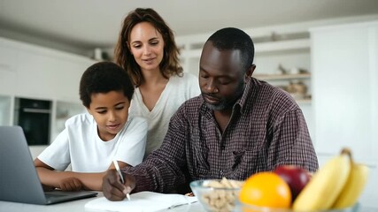  family learning moment in a bright modern kitchen. A Black father helps his teenage son with homework at the kitchen table, while the red-haired mother stands 