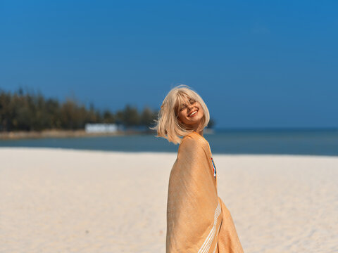 Smiling woman wrapped in orange shawl on sunny beach with blue sky, carefree and joyful, summer vacation and holiday mood.