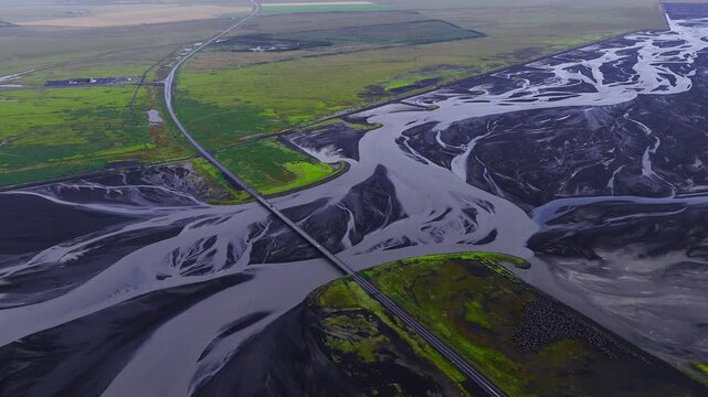 Aerial view shows a straight road and bridge across braided glacial channels on black volcanic outwash plains in south Iceland, with mossy islands in soft daylight.