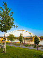 Naklejka premium White arch bridge over Danube River in Bratislava Slovakia. Scenic modern bridge crossing the Danube with green park foreground and blue sky