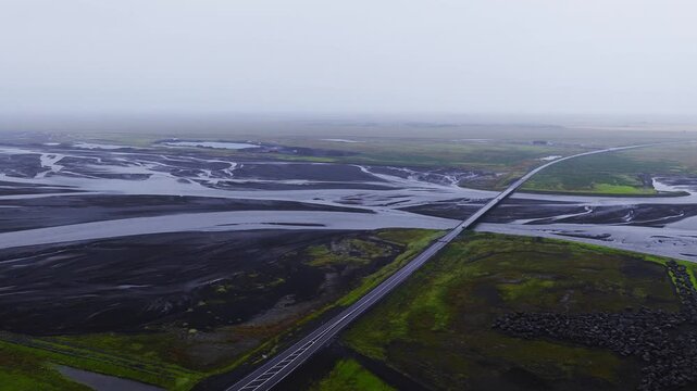 Aerial camera glides over Ring Road bridge on Iceland south coast as vehicles cross above braided glacial channels, dark sandbanks, green patches, and rock embankments under overcast light.
