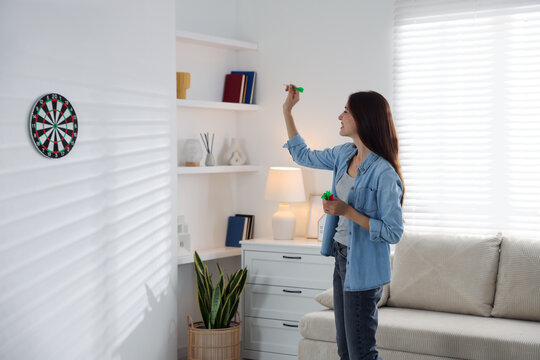 Happy woman with darts aiming at dartboard indoors