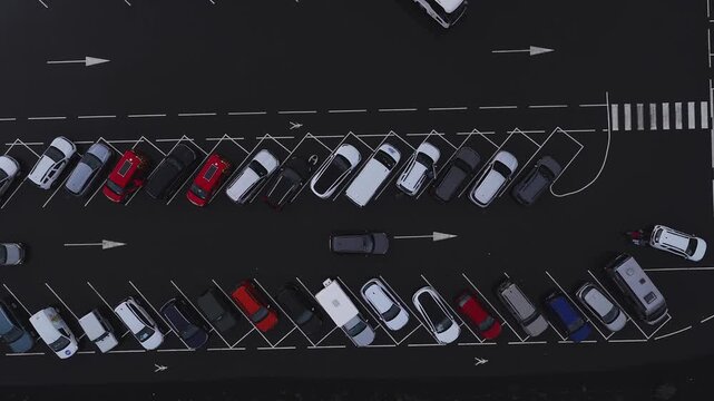 Top down aerial view of an airport parking lot in Iceland. Cars line two rows, a few move in the center lane. White arrows, zebra crossing, and wheelchair symbols stand out.
