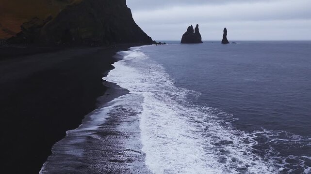 Aerial view shows Reynisdrangar sea stacks at Reynisfjara near Vik, Iceland. Foamy waves wash black sand by basalt cliffs, tiny visitors add scale under overcast light.