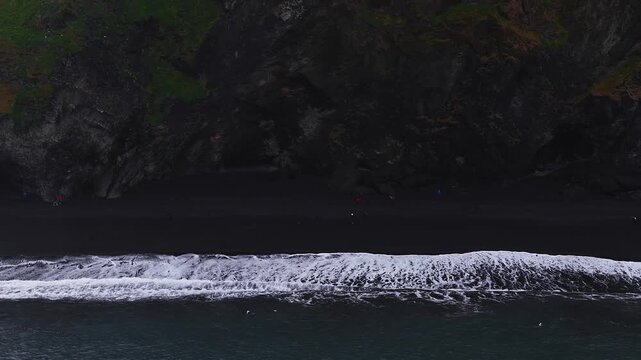 Aerial view shows Reynisfjara in Vik, Iceland as waves roll onto black sand below basalt cliffs. Visitors and seabirds move along the shore in overcast daylight.