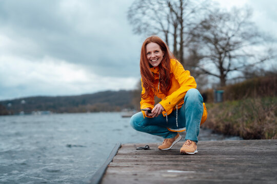Smiling redhead woman squatting on pier by lake