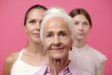 Portrait of senior Caucasian woman standing in front with middle aged Caucasian woman and teenage Caucasian girl in background, all looking directly at camera against pink backdrop