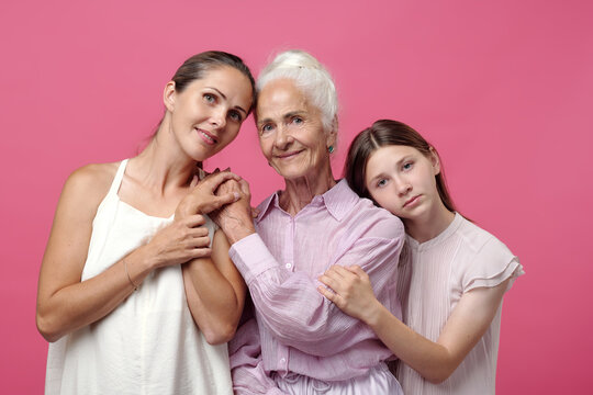 Caucasian middle aged woman, senior woman, and preteen girl standing close together holding hands and embracing, showing multigenerational family connection against pink background