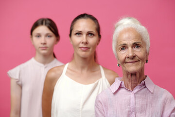 Caucasian senior woman standing in foreground looking at camera with slight smile, middle aged Caucasian woman and Caucasian teenage girl standing behind against pink background