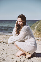 Young woman in white knitted dress crouching on Baltic Sea beach in soft daylight